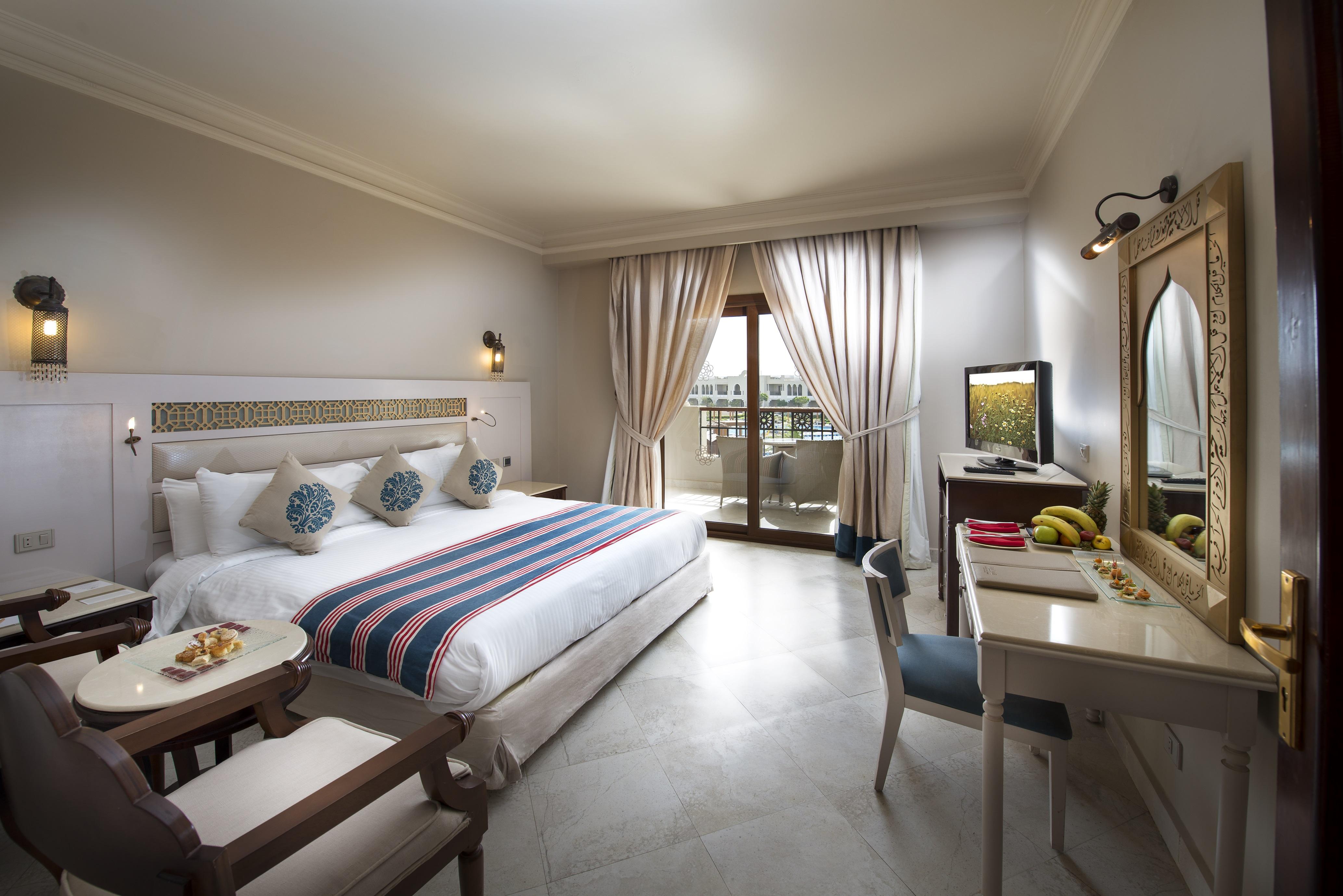 Sunrise Arabian Beach Resort Sharm El-Sheikh Exterior foto The photo depicts a hotel room that appears stylish and well-furnished. It features a large bed with decorative pillows and a colorful blanket. There is a small seating area with armchairs and a round table, as well as a work desk with a chair. A tel