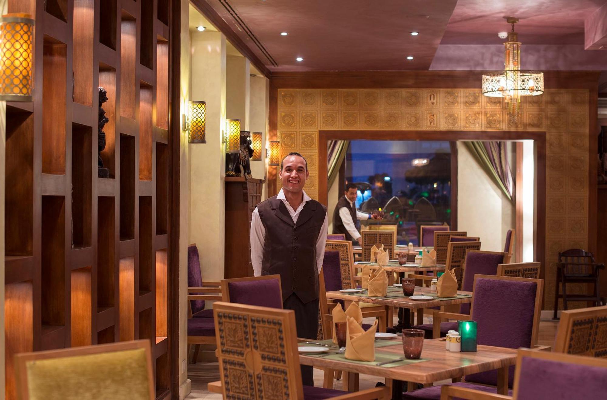 Sunrise Arabian Beach Resort Sharm El-Sheikh Exterior foto The photo depicts the interior of a restaurant featuring a staff member standing in the foreground. The restaurant has a warm and inviting ambiance, characterized by wooden accents, stylish lighting, and neatly arranged tables. The seating includes p
